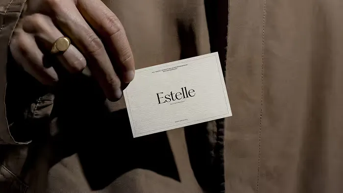 Hand holding a white business card with the name 'Estelle' printed in black serif font against a brown fabric background.