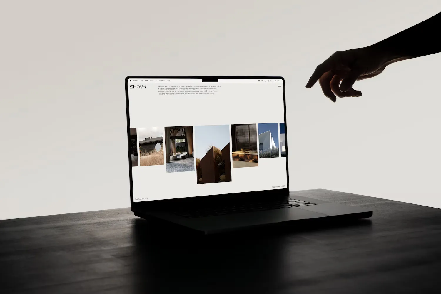 Laptop on a dark wooden table displaying an architecture portfolio website with a hand reaching toward the screen from the right.