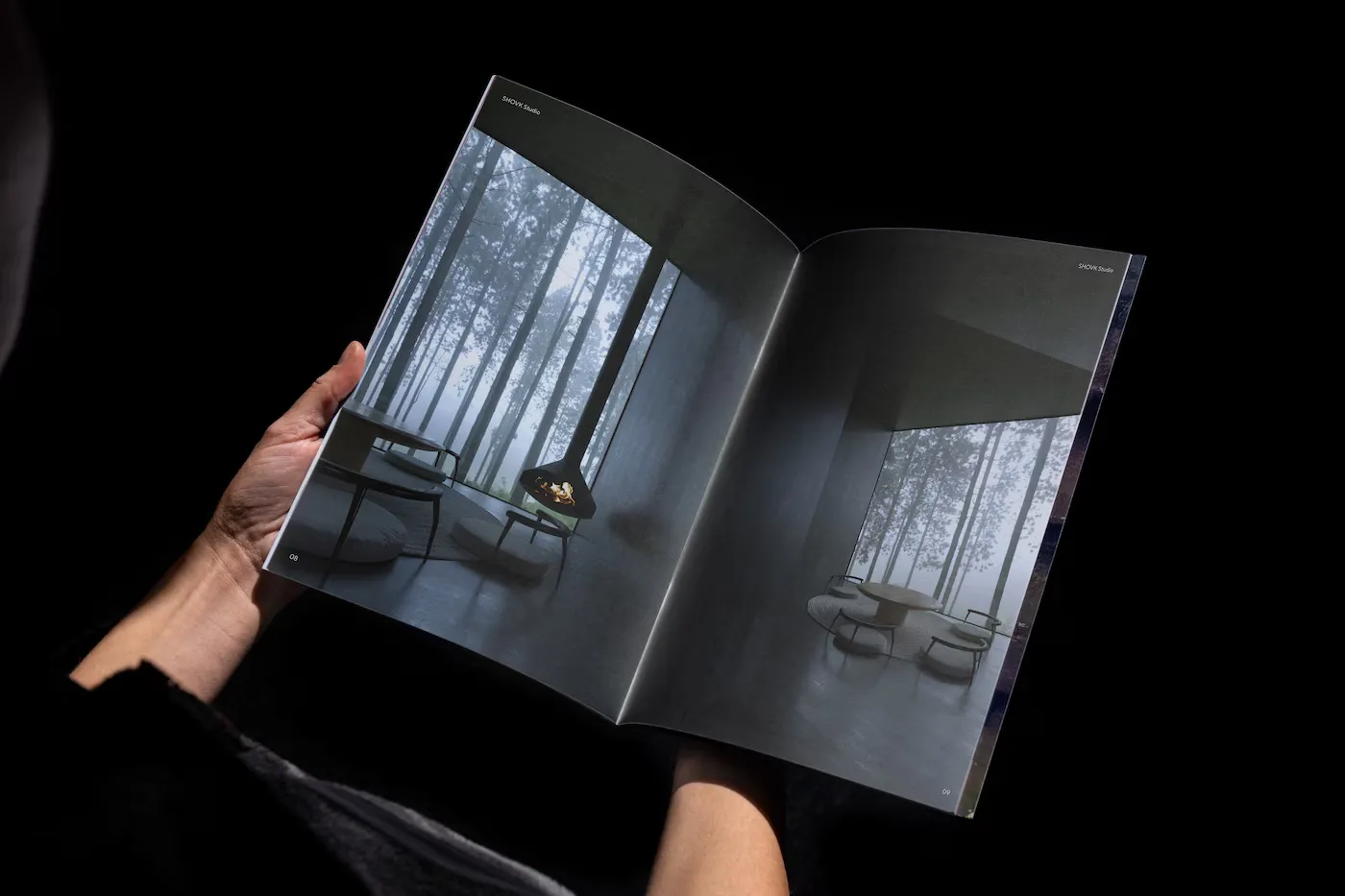 Open magazine held by hands showing modern interior designs with large windows revealing tall trees outside.