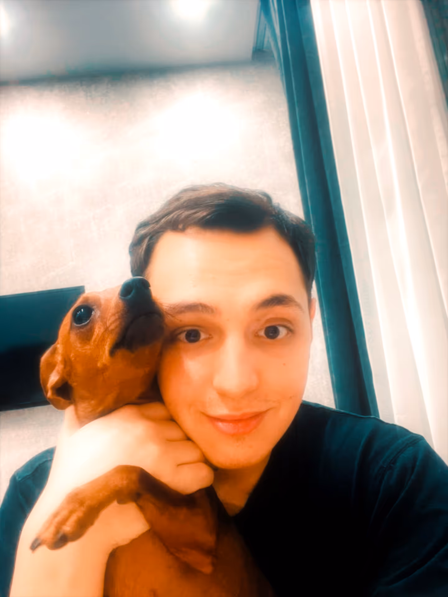 Young man smiling and holding a small brown dog close to his face indoors.