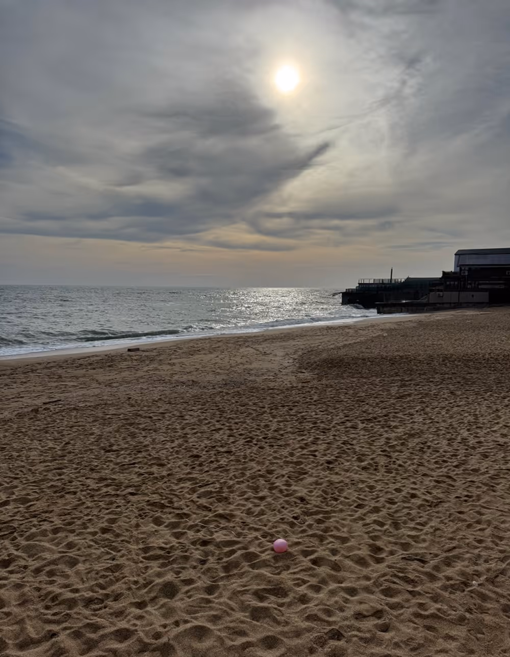 Sun partially covered by clouds over a calm sea with a sandy beach in the foreground and a small pink ball on the sand.