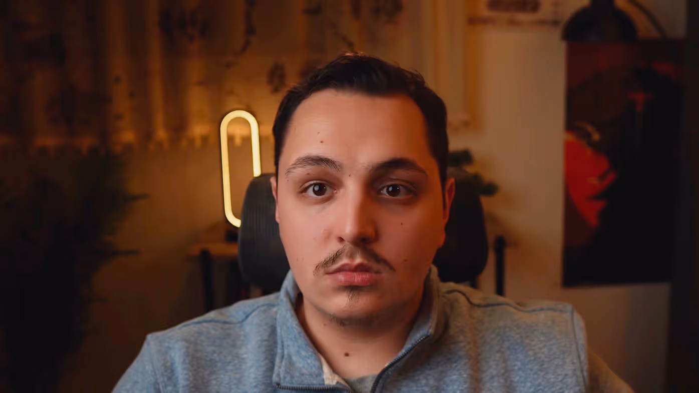 Young man with short dark hair and mustache wearing a gray zip-up sweater, sitting indoors with soft warm lighting.