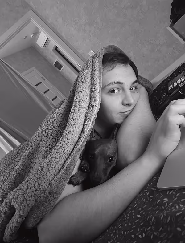 Person lying on a bed with a blanket over their head, holding a small dog, in a cozy room setting, black and white filter.